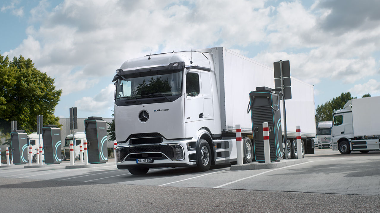 Ein weißer eActros 600 mit ProCabin von Mercedes-Benz Trucks mit einem weißen Anhänger steht auf einem Parkplatz und wird an einer Ladestation aufgeladen. Um den Lkw herum sind mehrere weitere Ladestationen und Absperrpfosten zu sehen. Im Hintergrund stehen weitere weiße Lastwagen, und am linken Bildrand sind grüne Bäume unter einem teilweise bewölkten Himmel zu erkennen.