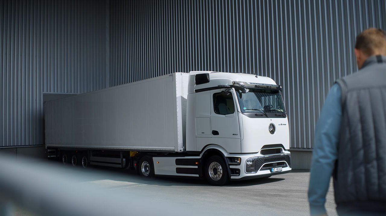 Ein weißer eActros 600 mit ProCabin von Mercedes-Benz Trucks mit einem langen, weißen Anhänger steht auf einem asphaltierten Gelände vor einer grauen Wellblechwand. Der Lkw ist von der Seite zu sehen, wobei die Front leicht nach links zeigt. Im Vordergrund, mit dem Rücken zum Betrachter, steht eine Person in einer blauen Jacke und einer grauen Weste, die auf den Lastwagen blickt. Das Licht fällt von oben auf die Szene.