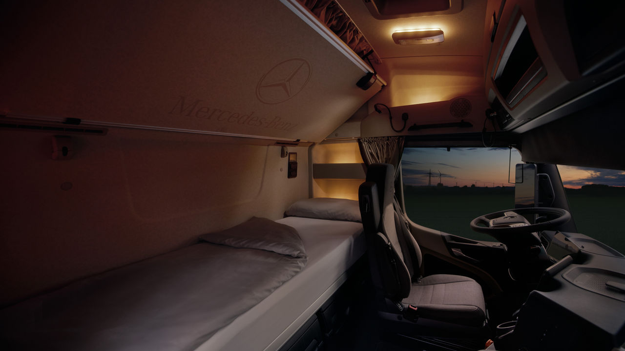 Das Bild zeigt das Innere der Kabine eines eActros von Mercedes-Benz Trucks bei Dämmerung. Links ist ein ordentlich gemachtes Bett mit hellgrauer Bettwäsche zu sehen, darüber an der Decke das Mercedes-Benz Logo und der Schriftzug "Mercedes-Benz". Rechts davon befindet sich der Fahrersitz und das Lenkrad, während durch die Windschutzscheibe ein dunkler Himmel mit den Silhouetten von Windrädern und einer weiten Landschaft zu erkennen ist.