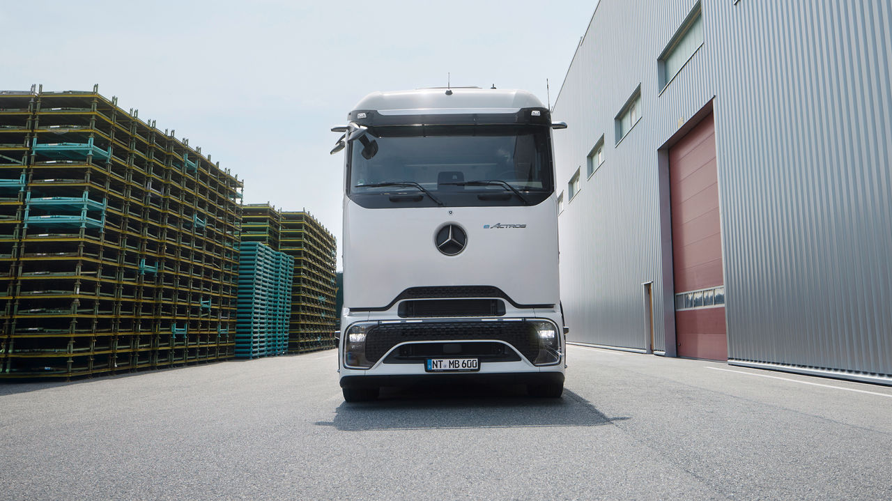 Ein eActros 600 mit ProCabin von Mercedes-Benz Trucks steht frontal auf einem asphaltierten Gelände. Links im Bild sind hohe Stapel von gelben und blauen Paletten zu sehen, während rechts ein großes, modernes Gebäude mit einer grauen Wellblechfassade und einem roten Tor steht. Der Himmel ist hell und leicht bewölkt.