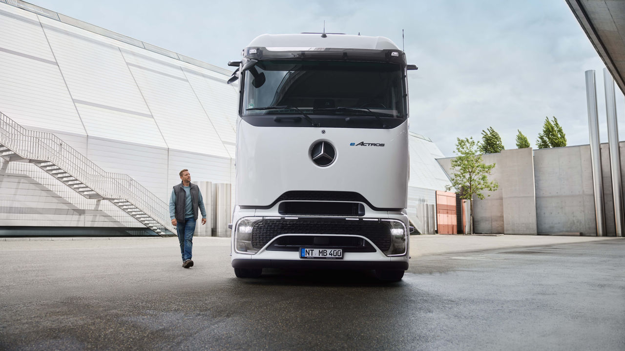 Ein Mann in Arbeitskleidung geht auf einer asphaltierten Fläche, während im Hintergrund ein weißer eActros 400 mit ProCabin von Mercedes-Benz Trucks parkt. Links sind ein modernes Gebäude mit einer langen Außentreppe zu sehen und rechts Bäume und weitere Gebäudestrukturen.
