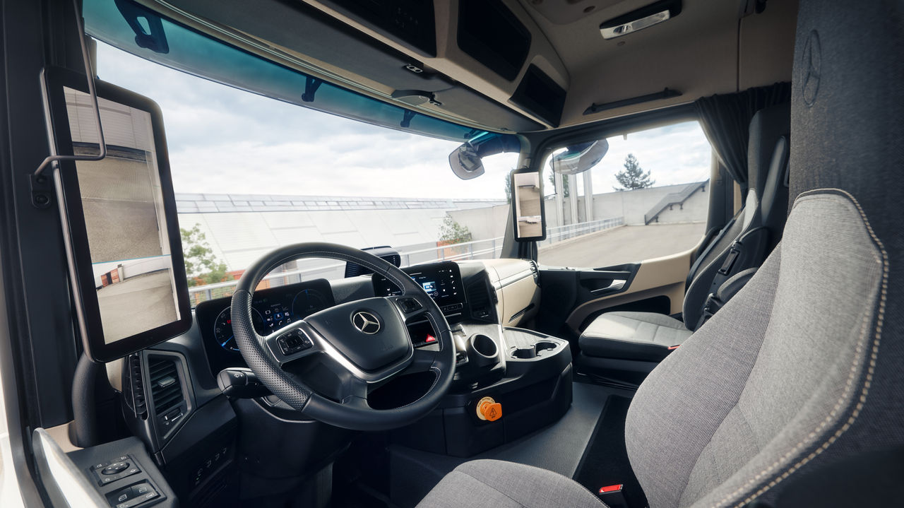 Das Bild zeigt das Innere des eActros von Mercedes-Benz Trucks aus der Perspektive des Beifahrersitzes. Man sieht das Lenkrad mit dem Mercedes-Stern, das Armaturenbrett mit digitalen Anzeigen und Bildschirmen, sowie die bequemen Sitze in hellgrauem Stoff.
