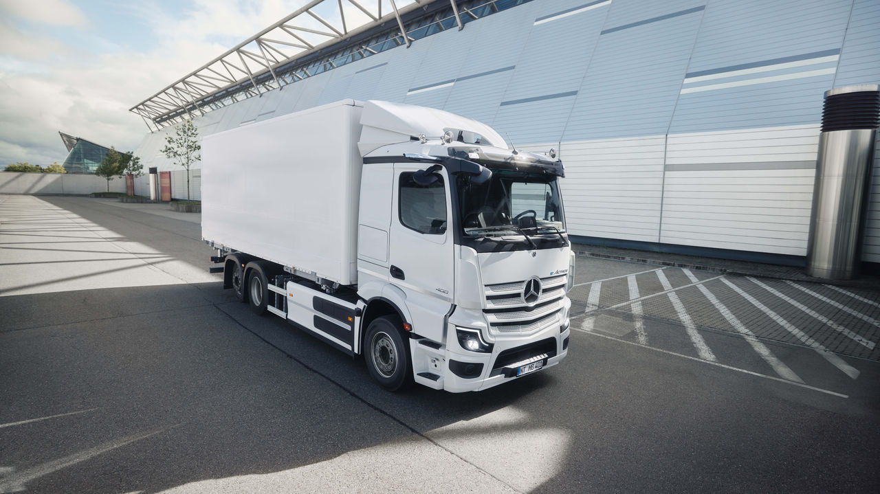 Ein weißer eActros 400 von Mercedes-Benz Trucks mit einem Kofferaufbau steht auf einem asphaltierten Parkplatz vor einem modernen Gebäude mit einer Glasfassade. Das Sonnenlicht wirft lange Schatten auf den Boden, was auf einen klaren Tag hindeutet.